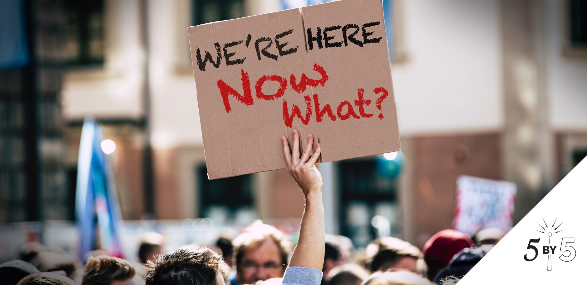 man's arm above crowd holding sign "We're Here. Now What?"